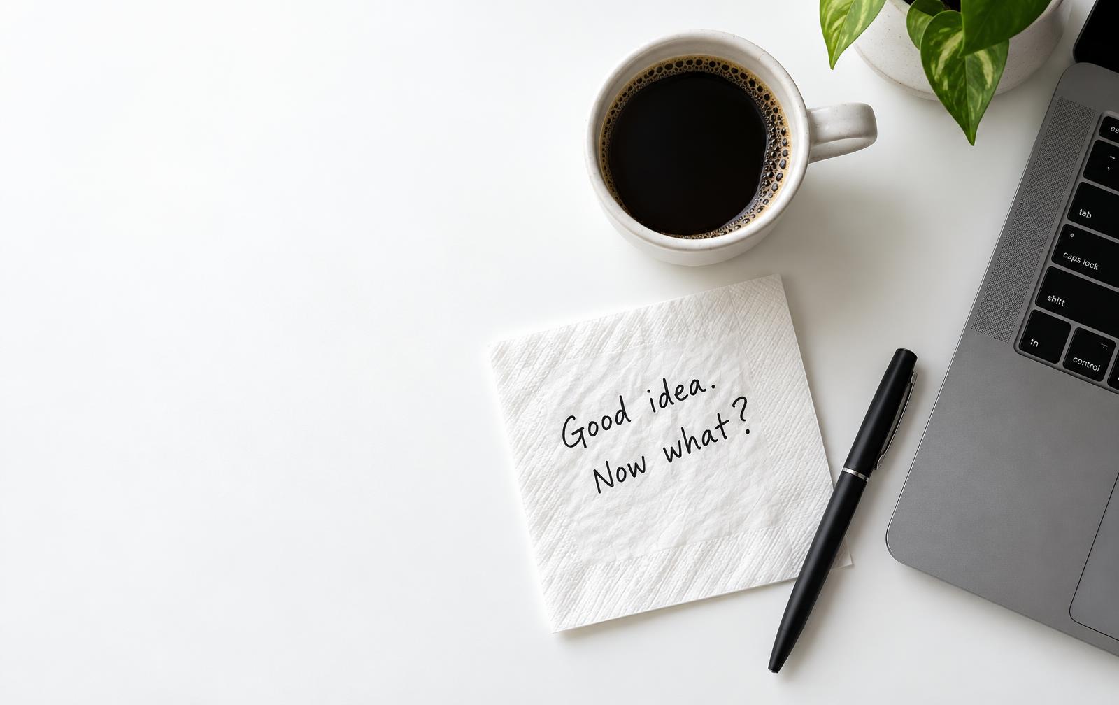 A napkin reading Good idea. Now what? beside a coffee and a laptop
